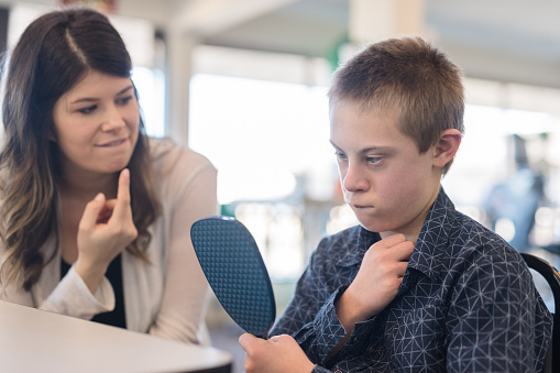 A female speech therapist works with a handsome elementary-age boy in a clinic setting. He is holding a mirror and watching himself pronounce each syllable.