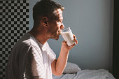 Adult man illuminated with sunlight through blinded window, drinking glass of milk early in the morning, after waking up