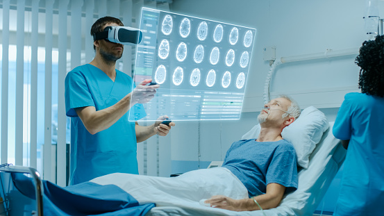 Doctor Wearing Virtual Reality Headset and Holding Joysticks Cures Senior Patient who is Lying in Bed. He Uses Augmented Reality Interface, Looks at Brain Scans and Medical History. Nurse Does Checkup
