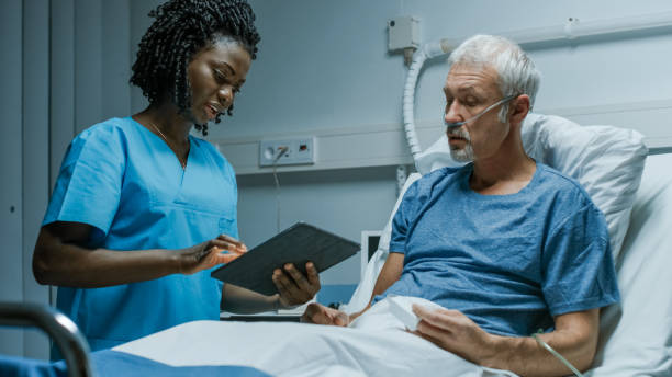 in the hospital, senior patient lying in the bed talking to a nurse who is holding tablet computer showing him information. in the technologically advanced hospital ward. - hospital patient bed nurse imagens e fotografias de stock