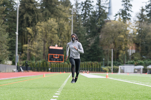 An African American female track athlete trains with a high altitude training mask at a stadium. She is running sprint drills on the field while she wears it.
