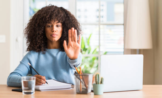 istock African american woman studying at home using a computer laptop with open hand doing stop sign with serious and confident expression, defense gesture 1043180138