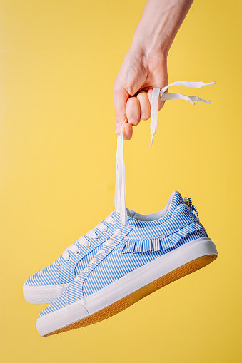 female hand holds blue striped sneakers for laces on yellow background. Concept photo with sneakers and pastel colors