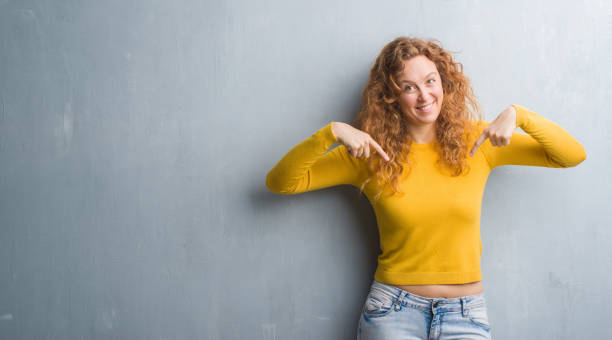 istock Young redhead woman over grey grunge wall looking confident with smile on face, pointing oneself with fingers proud and happy. 1043233262
