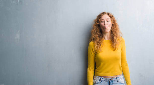 istock Young redhead woman over grey grunge wall making fish face with lips, crazy and comical gesture. Funny expression. 1043233740