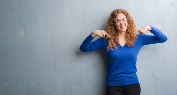 istock Young redhead woman over grey grunge wall looking confident with smile on face, pointing oneself with fingers proud and happy. 1043245362