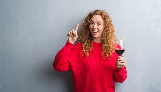 Young redhead woman over grey grunge wall drinking a glass of wine surprised with an idea or question pointing finger with happy face, number one
