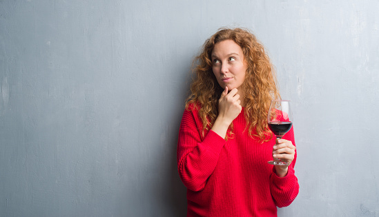 Young redhead woman over grey grunge wall drinking a glass of wine serious face thinking about question, very confused idea