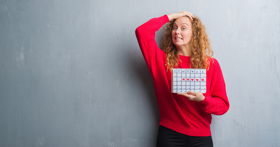 Young redhead woman over grey grunge wall holding period calendar stressed with hand on head, shocked with shame and surprise face, angry and frustrated. Fear and upset for mistake.