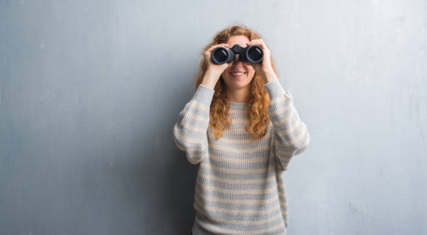 Young redhead woman over grey grunge wall looking through binoculars with a happy face standing and smiling with a confident smile showing teeth