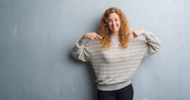 istock Young redhead woman over grey grunge wall looking confident with smile on face, pointing oneself with fingers proud and happy. 1043255830