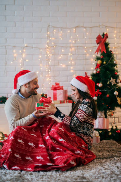 istock Presents time ! Young happy couple sharing new years presents while sitting on floor covered with blanket. Colorful christmas decoration in background. 1043262936