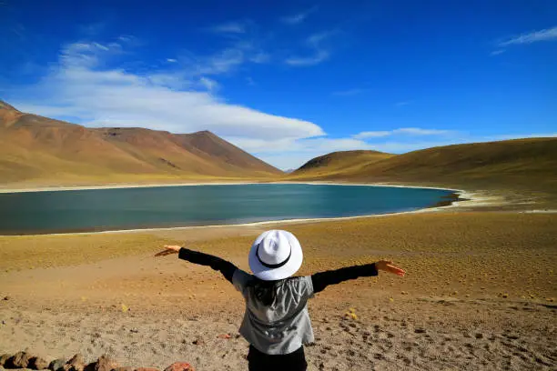 Female tourist raising her arms admiring the amazing deep blue lagoon Laguna Miniques, located in the altiplano of Antofagasta region, Chile