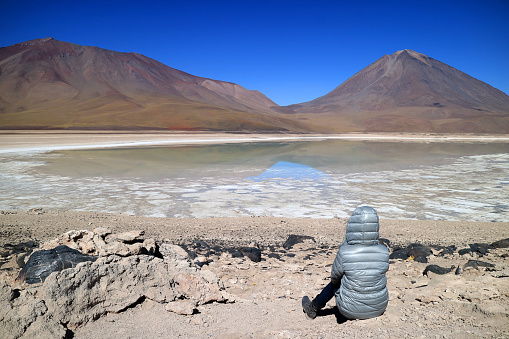 Woman sitting on the shore waiting for the green water effect of Laguna Verde or the Green Lake with Lincancabur Volcano in backdrop, Potosi, Bolivia