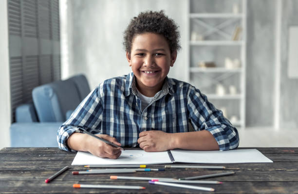 Afro American boy Happy Afro American boy is drawing using colored pencils, looking at camera and smiling while sitting at the table at home sad african child drawings stock pictures, royalty-free photos & images