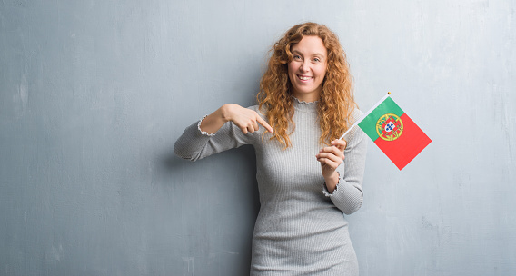 Young redhead woman over grey grunge wall holding flag of Portugal with surprise face pointing finger to himself