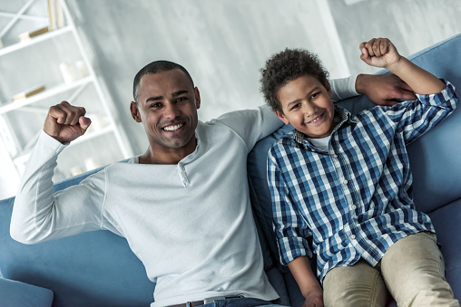 Happy Afro American father and son in casual clothes are raising their fists, looking at camera and smiling while sitting on couch at home