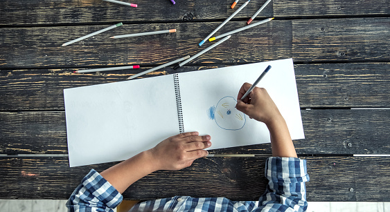 Top view of Afro American boy drawing using colored pencils, on wooden table, cropped