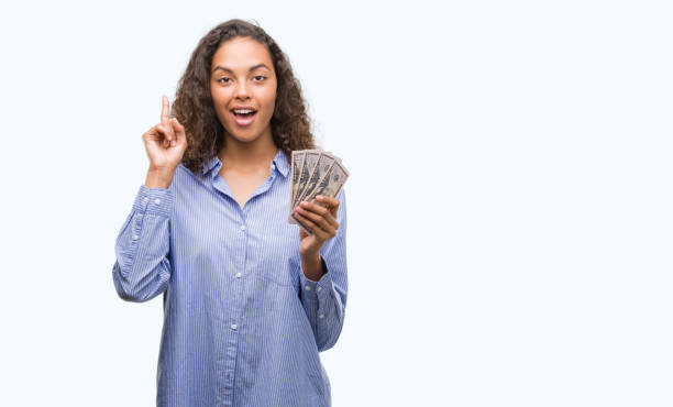 istock Young hispanic woman holding dollars surprised with an idea or question pointing finger with happy face, number one 1043281642