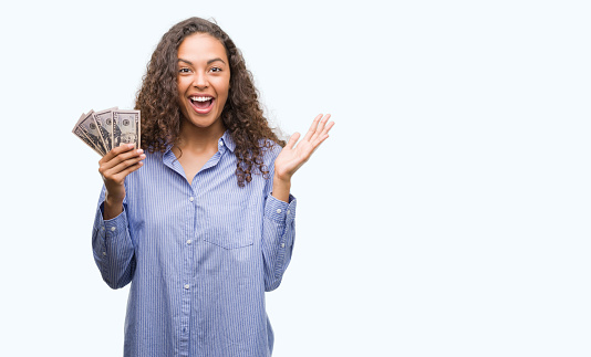 Young hispanic woman holding dollars very happy and excited, winner expression celebrating victory screaming with big smile and raised hands