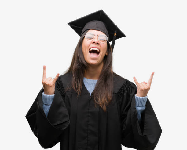 Young hispanic woman wearing graduated cap and uniform shouting with crazy expression doing rock symbol with hands up. Music star. Heavy concept.