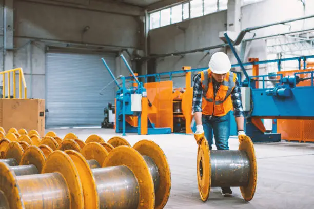 Skilled young heavy industry engineering worker man loading and working with punching bobbin metal reel cable drum reel wire spools with safety workwear and moving a massive metal construction object in warehouse of factory. XXXL size
