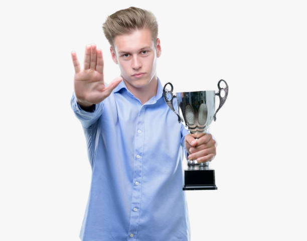 istock Young handsome blond man holding a trophy with open hand doing stop sign with serious and confident expression, defense gesture 1043302938