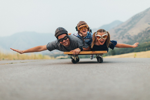 Two young children, a boy and a girl, are imagining flying into the sky while riding on a press cart with their father. They have their arms spread out like wings and ready to use imagination in being like an airplane and piloting the airplane into the sky. They love spending time together as a family. They are wearing flying goggles on a rural road in Utah, USA.