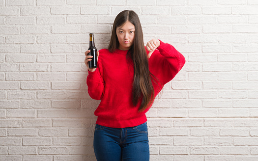 Young Chinese woman over brick wall drinking beer with angry face, negative sign showing dislike with thumbs down, rejection concept