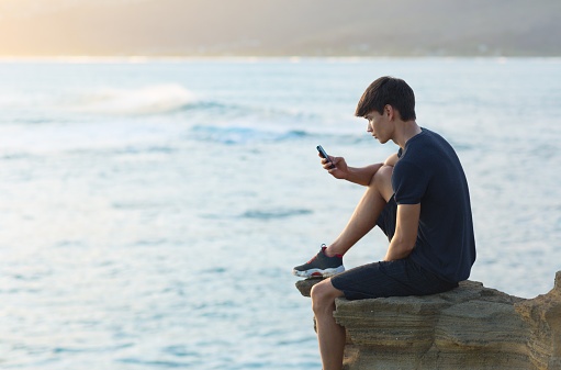 Young Caucasian man sitting at the edge of the rocks next to the ocean texting on his phone.