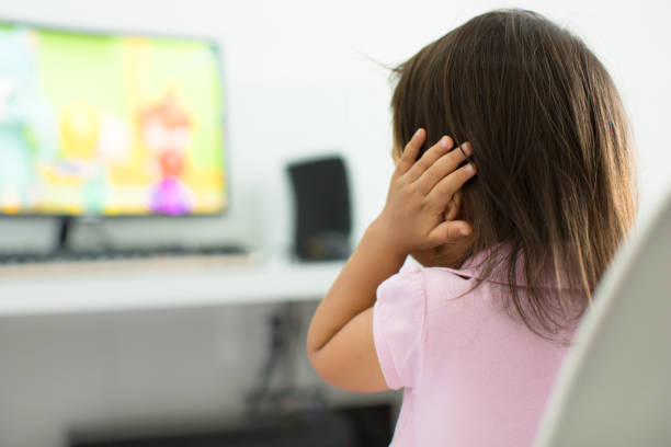 A terrified child, afraid of the loud sounds from the television. Autism. A child watching tv holding her ears because she is afraid of the sound; Child behavior theme asian kids watching tv stock pictures, royalty-free photos & images
