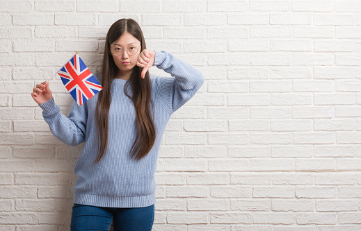 Young Chinese woman over brick wall holding flag of England with angry face, negative sign showing dislike with thumbs down, rejection concept