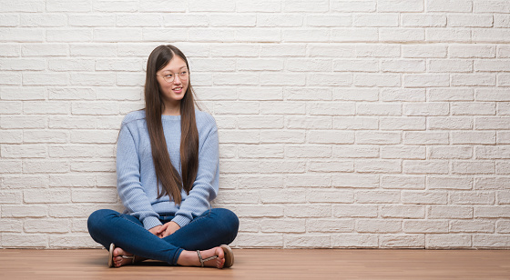 Young Chinese woman sitting on the floor over brick wall smiling looking side and staring away thinking.