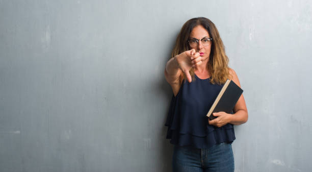 middle age hispanic woman standing over grey grunge wall holding a book with angry face, negative sign showing dislike with thumbs down, rejection concept - women professor mature adult human face imagens e fotografias de stock