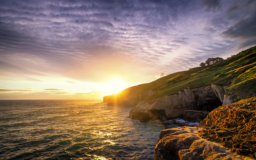 An epic sunset with dramatic burning clouds in Tunnel Beach of Dunedin, New Zealand. One can enjoy ocean view, cliff, rolling hills, sunset, coves, rock formation in this famous tourist attraction.