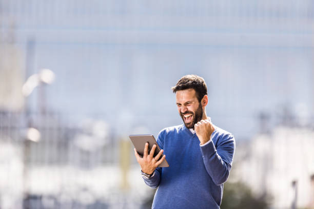 Cheerful businessman using touchpad and screaming of joy in the city. Joyful businessman reading a great news over digital tablet and celebrating his success on the street. Copy space. businessman happiness outdoors cheerful stock pictures, royalty-free photos & images
