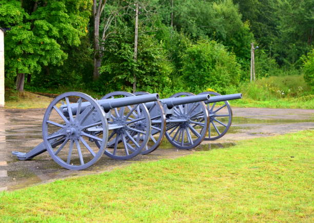 istock Three small cannons arranged in a straight line standing on an asphalt square near a freshly mown grass seen right after a strong rain, with puddles everywhere and water dripping from the objects 1043497362