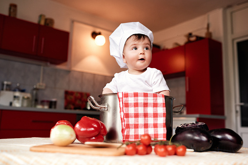 funny baby girl wearing chef's hat, posing inside pot, surrounded by begetables.