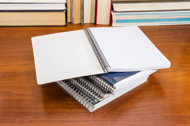 Open blank exercise book with pages of squared paper and wire spiral binding lies on other notebooks on the wooden table against of books