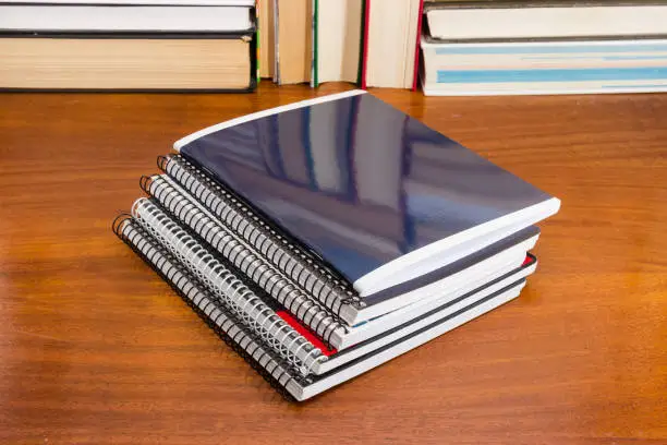 Stack of the different exercise books with wire spiral binding on the wooden table against of books
