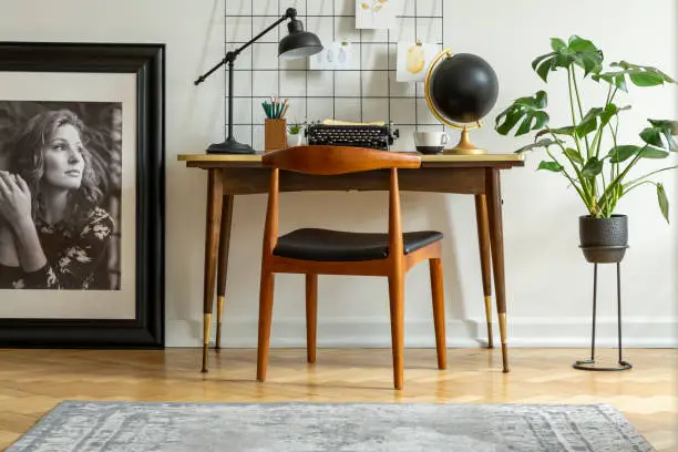 Photo of Mid-century modern chair with leather seat by a desk with an industrial lamp and a retro typewriter in a white home office interior