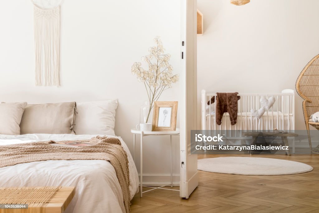 Real photo of white bedroom interior with bedside table with plant, poster and mug and open door to baby room with rug and white crib Herringbone Stock Photo