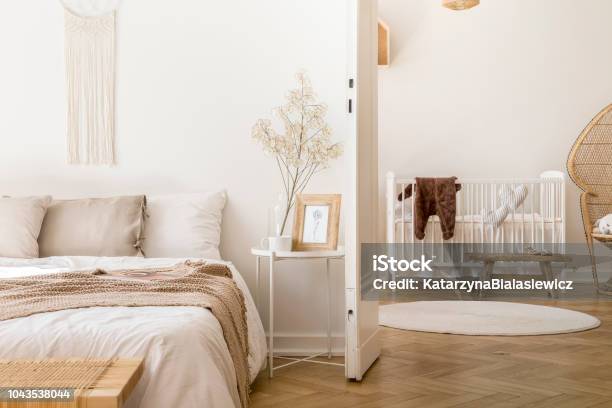 Real Photo Of White Bedroom Interior With Bedside Table With Plant Poster And Mug And Open Door To Baby Room With Rug And White Crib Stock Photo - Download Image Now