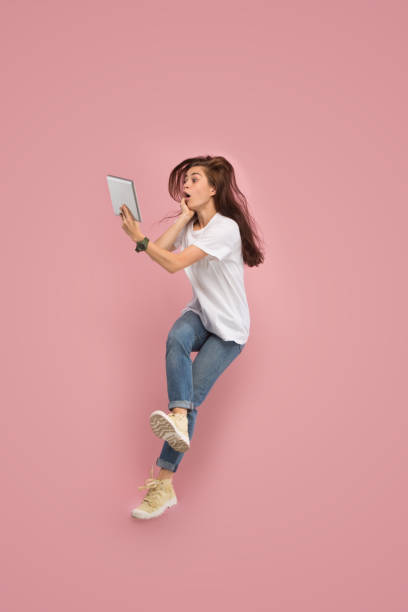 istock Image of young woman over pink background using laptop computer or tablet gadget while jumping. 1043562022