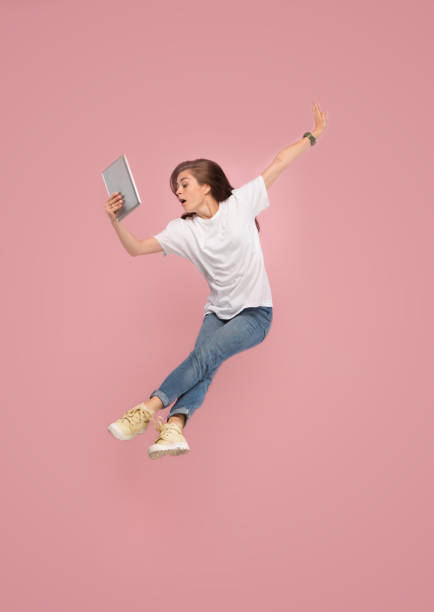 istock Image of young woman over pink background using laptop computer or tablet gadget while jumping. 1043562124