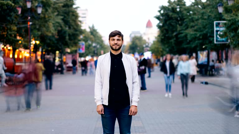 Zoom out time-lapse of confident young man looking at camera standing in city center in pedestrian street while men and women are walking around him in hurry.