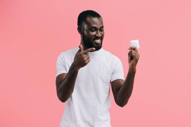 souriant jeune homme afro-américain pointant au préservatif isolé sur fond rose - condom photos et images de collection