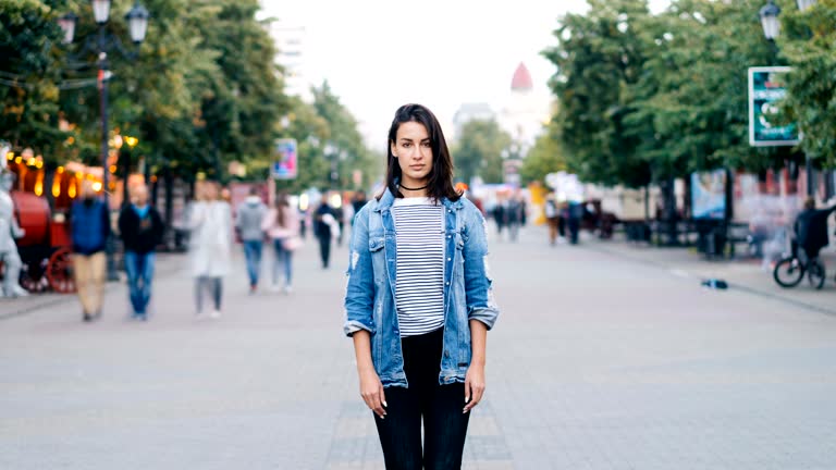 Zoom out time-lapse of beautiful lady with dark hair standing alone among crowd of people and looking at camera. Modern life, society and loneliness concept.