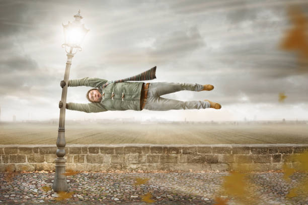 A man with a funny facial expression is holding on to a lamp post while a heavy storm ist blowing. The man is flying horizontally in mid air.