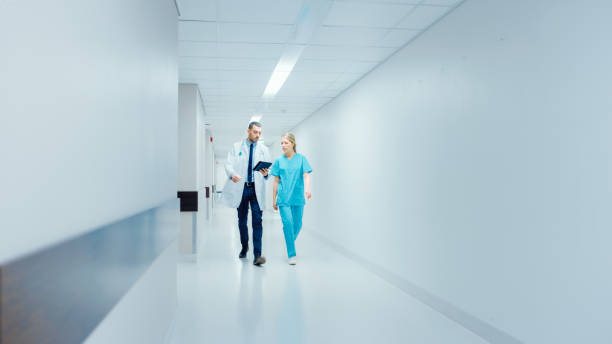 istock Surgeon and Female Doctor Walk Through Hospital Hallway in a Hurry while Using Digital Tablet and Talking about Patient's Health. Modern Bright Hospital with Professional Staff. 1043634370
